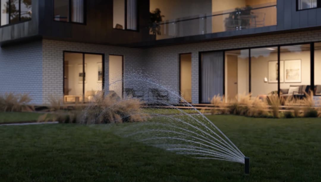Automatic sprinkler system watering lush lawn in modern backyard at dusk.
